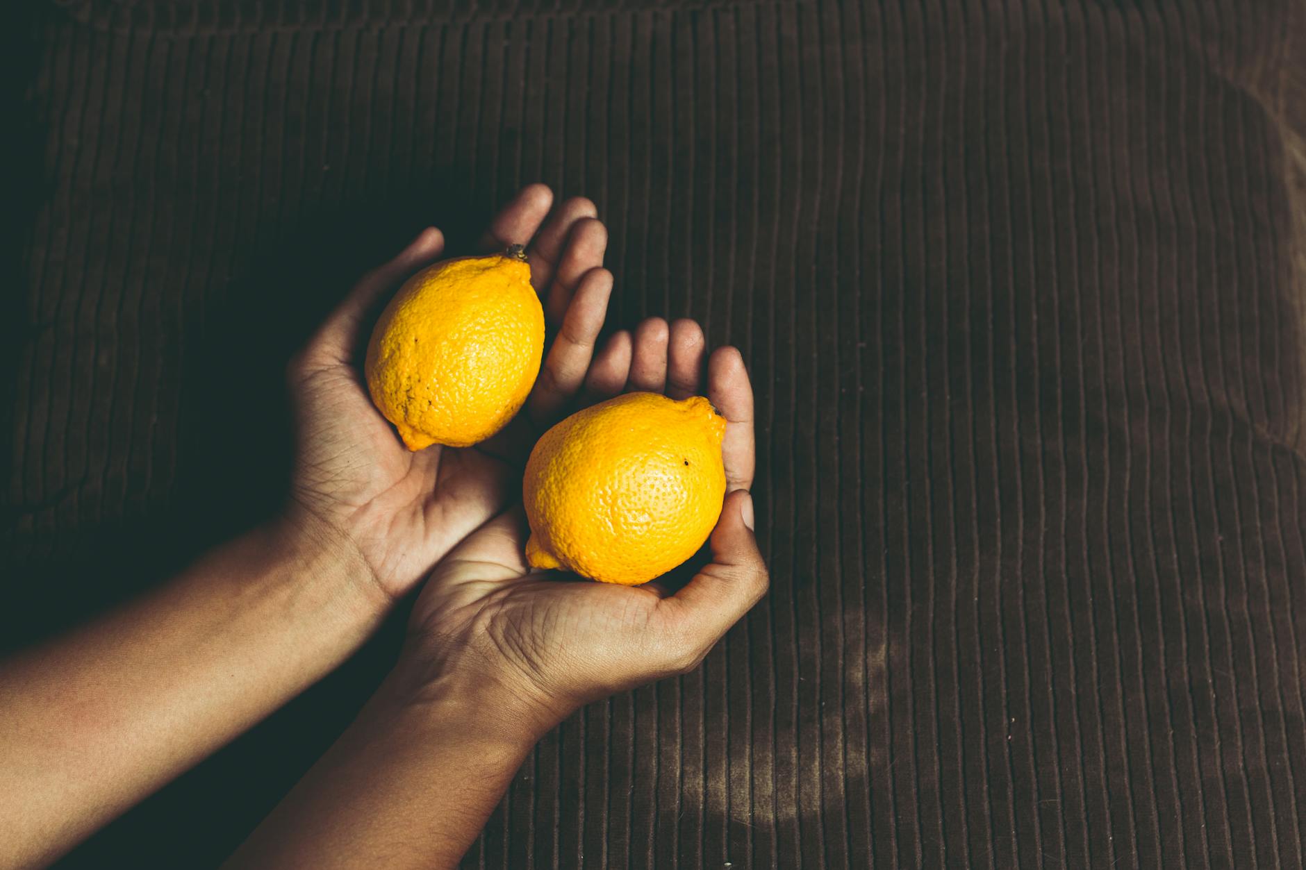Hand holding a fresh lemon on a soft pink background with additional lemons nearby, representing refreshed sensation